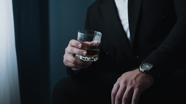 A man in a suit holding a glass of whiskey, with a focus on the hand and the watch, against a dark, blurred background, suggesting sophistication.