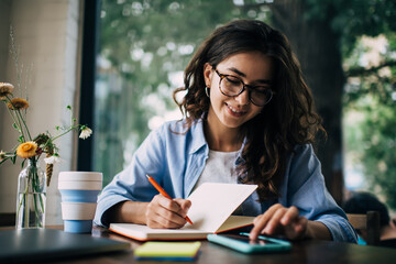 Cheerful woman taking notes and browsing smartphone in cafe