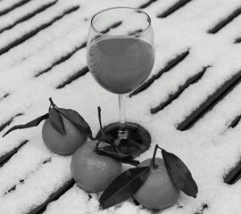 Glass of organic fresh squeezed mandarin juice on snow covered table in garden and tangerine fruits. Healthy eating lifestyle background. Energy shot before winter sport activities. Black white photo.