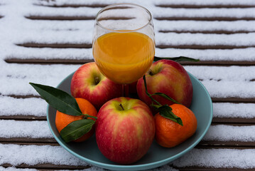 Glass of organic fresh squeezed fruit juice on a snow covered table in the garden and tangerines with apples in a bowl. Healthy eating lifestyle background. Energy shot before winter sport activities.