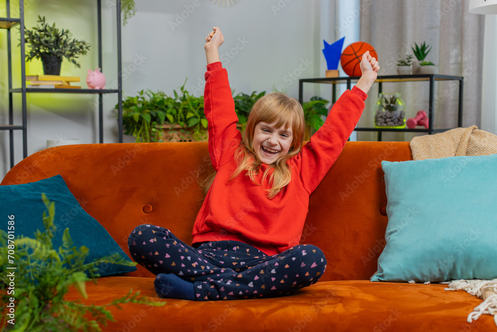 Wall mural Happy child girl shouting, celebrating success, winning, goal achievement good news, lottery luck, victory, watching TV championship at modern home apartment. Teen kid in living room sitting on couch