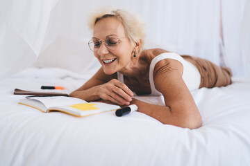 Happy aged woman reading notebook while lying on bed