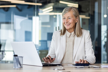 Senior businesswoman smiling while working at laptop in modern office setting, exuding confidence...