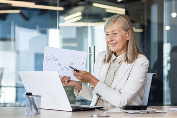 Confident senior businesswoman analyzes financial charts in a modern office, portraying professionalism and concentration.