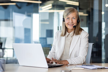 Professional middle-aged woman with headset working on laptop in modern office, displaying customer...