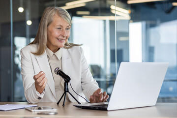 Confident senior businesswoman giving a presentation, speaking into a microphone at her laptop in a...