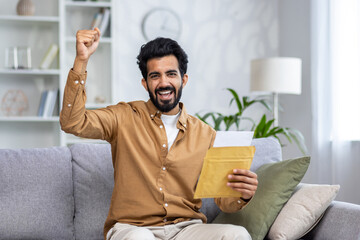 Joyful man with a beard celebrating while holding an envelope, sitting on a sofa at home, conveying...
