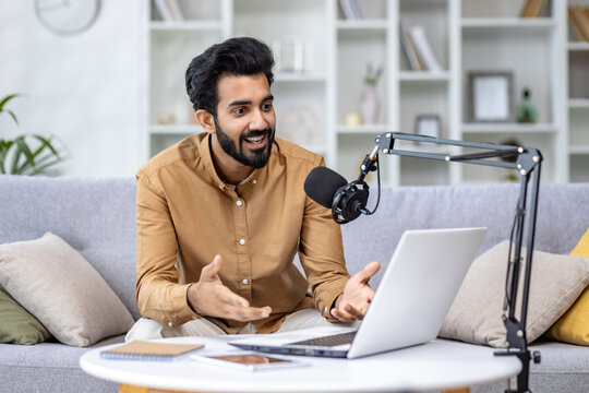 Enthusiastic man podcasting with laptop and microphone at home, expressing positivity and engagement in a cozy setting.