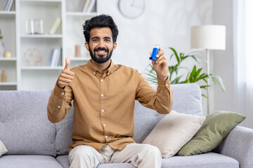 Smiling man sitting on couch giving thumbs up while holding asthma inhaler, promoting health and...