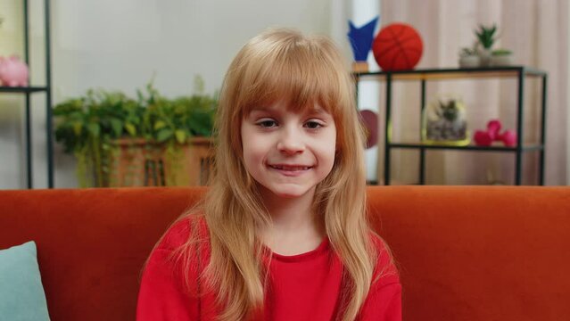Close-up portrait of happy smiling cheerful Caucasian preteen school girl 8 years old. Young lovely blonde little child kid looking at camera at home play living room apartment sitting on orange couch