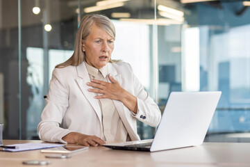 Senior woman in business suit sitting at desk in office in front of laptop and holding hand to chest, having panic attack, heart attack, shocked by business affairs. - obrazy, fototapety, plakaty