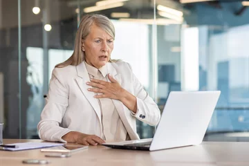 Fotobehang Senior woman in business suit sitting at desk in office in front of laptop and holding hand to chest, having panic attack, heart attack, shocked by business affairs. © Liubomir