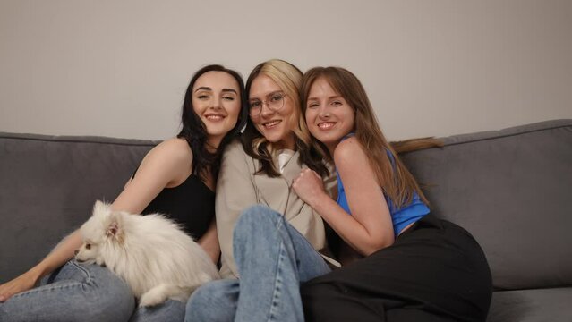 Three girlfriends are taking photos, sitting on the sofa. The girl sets the self-timer on the camera and sits down with her friends on the sofa. The girls are sitting cuddling to each other, smiling