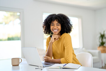Happy young African American lady sitting at home table and looking at camera while hybrid working...