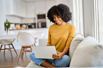 Obraz na płótnie Canvas Happy young African American woman sitting on couch at home using laptop. Smiling ethnic lady relaxing on sofa looking at computer technology device browsing web buying online doing shopping.