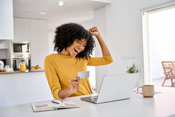 Happy excited young African woman customer winner holding credit card using laptop computer in kitchen celebrating money win receiving cashback getting online bonus for shopping sitting at home. - Powered by Adobe