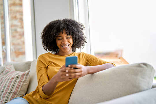 Smiling young African woman using cell phone relaxing on couch at home. Happy lady sitting on sofa looking at smartphone holding mobile cell phone in hands texting buying online in ecommerce shop.