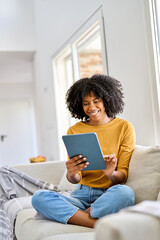 Happy young African American woman sitting on couch at home using digital tablet. Smiling girl...