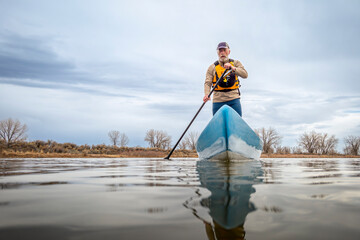 senior male paddler is paddling a stand up paddleboard on a calm lake in spring, frog perspective...