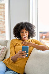 Excited young African American woman customer looking at mobile phone sitting on sofa at home....
