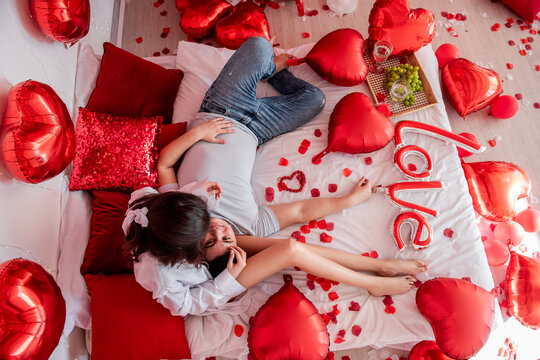 Top view of loving couple in casual clothes on white bed with red heart balloons. Woman hugs man, they look at each other, hold each others hands. Warm Valentines Day Party. Trendy interior design