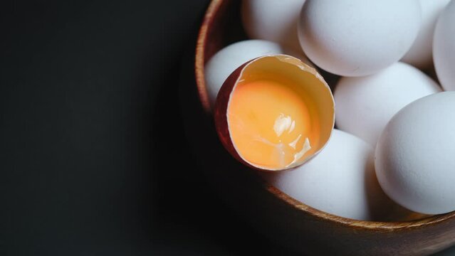 Broken egg with yolk on a background of eggs in a wooden bowl close-up on a black background