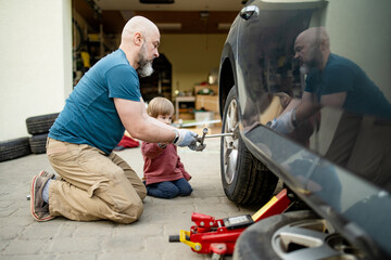 Cute toddler boy helping his father to change car wheels at their backyard. Father teaching his...
