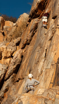 Far shot of a male rock climber on a steep rock face with a felame climber watching from the ground