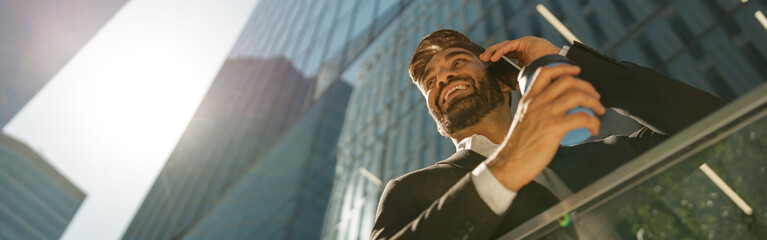 Smiling businessman is talking phone and drinking coffee standing on background of city skyscrapers