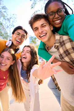 Vertical multiracial group of teenagers taking a selfie looking front camera laughing and having fun piggybacking together outdoor. Young students enjoying their free time with friends in street city
