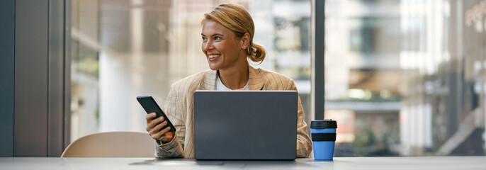 Stylish business woman use phone while sitting in modern coworking and working on laptop