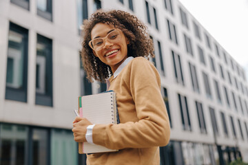 Smiling female freelancer standing with note pads on modern building backgrounds