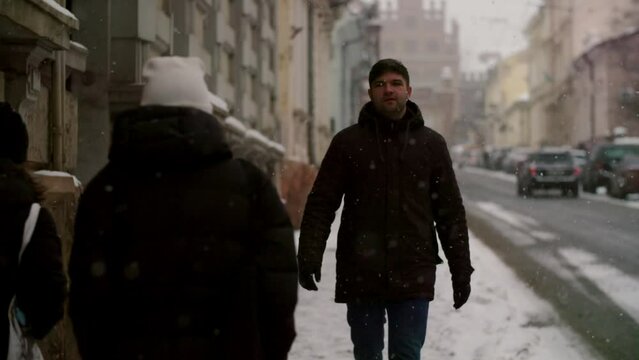 Slo mo young man tourist stroll city streets in icy weather outdoors. Male in winter jacket walking down the street exploring beautiful town. Snow fallling, cold weather and atmosphere