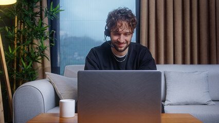 Smiling curly hair student man wear wireless headphone with microphone, looking at laptop, having video conference calling talk by webcam	