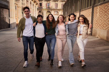 Multiracial group of smiling young friends walking hugging on street in historic city center. Happy tourists on school trip strolling embracing outdoors. Generation z tourism people enjoying together