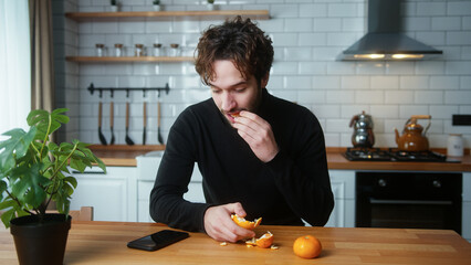Fototapeta na wymiar Young curly haired man sitting in the kitchen eating tangerines while talking on speakerphone on his mobile phone 