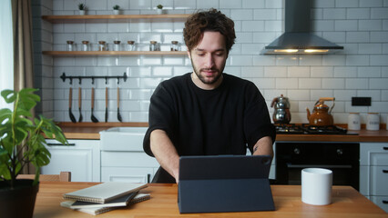 A young curly hair man sitting in kitchen, using app with tablet computer, surfing on internet	