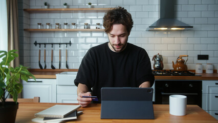 A curly hair man sitting in kitchen entering credit card number on tablet computer for makes secure...