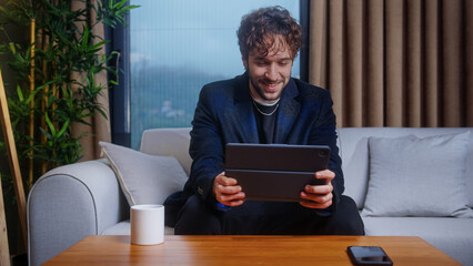 Happy curly hair man playing video game on a tablet while sitting on sofa in living room at home....