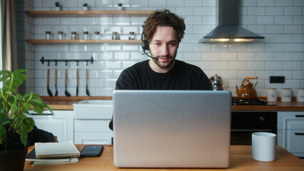 ,Smiling man sitting in kitchen at home wear wireless headset having video conference calling on laptop computer talk by webcam, learning study with online tutor	