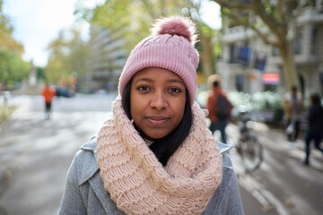 Beautiful serious latin woman standing in winter clothes smiling looking at camera holding shopping bag on the street in the city. Young female posing outdoors on holidays enjoys christmas vacation