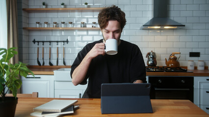 Young curly hair man sitting in kitchen, using app with tablet computer, surfing on internet	

