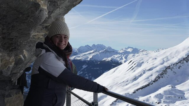 Woman posing for camera standing by handrail elevated metal support by cliffside with breathtaking view of mountains in background covered in snow