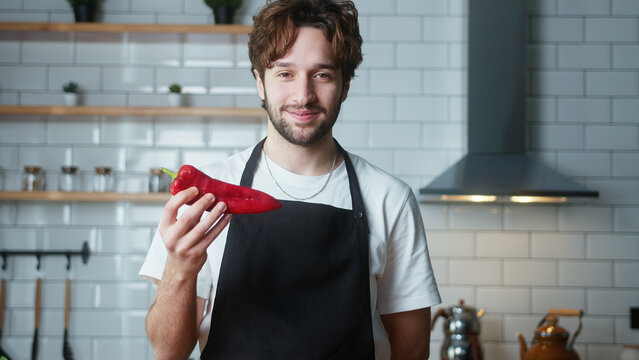 Young curly hair man in an apron holding red pepper in his hand recording video cooking lesson about vegetarian meal in modern kitchen