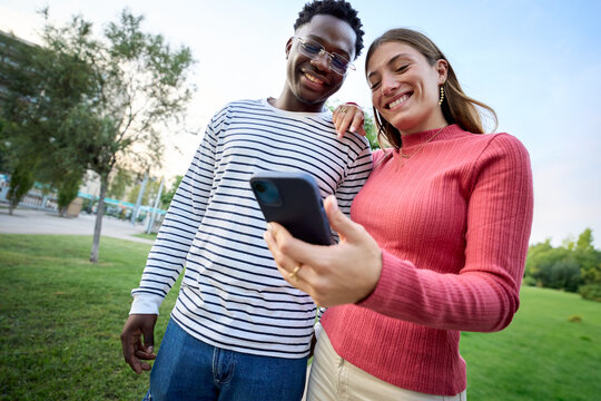Young multiracial couple smiling holding and looking at smart phone in their hands standing in a garden area. Two people cheerful and happily using mobile cell outdoors. Caucasian girl and African guy