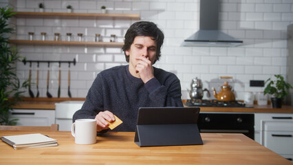 Sad man sitting in kitchen at home entering credit card number on tablet device for makes distant...