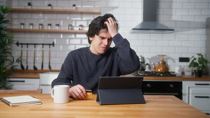 Young man sitting in kitchen at home entering credit card number on tablet device for makes distant goods purchase. Credit card is declined or out of limit, feeling sad and unhappy	
