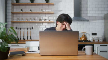 Young man sitting in the kitchen counter headache while using laptop. Tired young man get having...