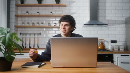 Happy man sitting in the kitchen entering credit card number on laptop for makes secure easy...