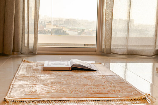 A muslim carpet set on the floor, beige brownish color with a mosque, curves and rectangle patterns. 
An opened Holy coran book placed on the rug in front of a balcony window. 